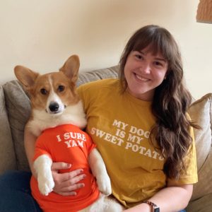 A woman holds a corgi dog in her lap while sitting on a couch. The woman has a mustard-colored shirt that reads “My dog is my sweet potato,” and the dog has an orange shirt that reads, “Yes I yam.”¬¬