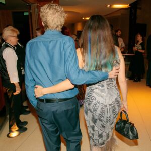 A young man with blonde hair and a young woman with brown and blue hair walk arm in arm away from the camera.